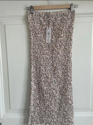 Blommig tubklänning från Design by Si - Supersöt tubklänning från Design by Si med blommigt mönster i beige, grönt och rosa på vit botten. Klänningen har smockad struktur och är figurnära. Perfekt för sommarens alla tillfällen! Storlek S