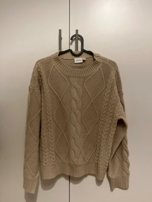 Beige kabelstickad tröja från Vila - Snygg beige kabelstickad tröja från Vila med grovt mönster och rund halsringning. Tröjan har långa ärmar och ribbade muddar vid ärmslut och nederkant. 