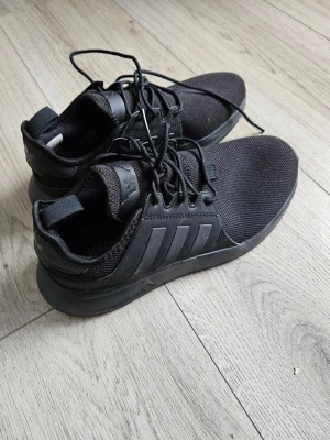 Svarta Adidas sneakers med mesh - Snygga helsvarta sneakers från Adidas med klassiska tre ränder på sidan. Ovandelen är i mesh och syntetmaterial för bra andningsförmåga och komfort. Skorna har lågt skaft och snörning framtill. Perfekta för en clean och sportig look.