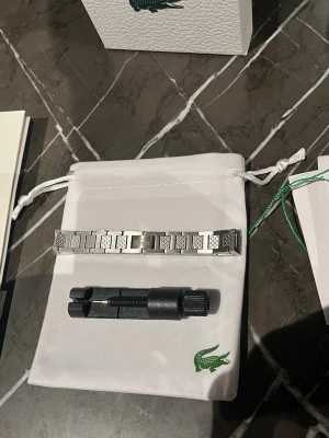 Lacoste silverfärgad klocklänk - Säljer en stilren silverfärgad klocklänk från Lacoste i rostfritt stål. Länken har ett rutigt mönster på länkarna och den klassiska Lacoste-loggan ingraverad på mitten. Perfekt för dig som vill byta ut eller uppgradera din klocka.