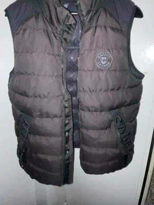 Svart dunväst från Zavetti Canada - Snygg svart dunväst från Zavetti Canada med broderad logga på bröstet och ryggen. Västen har hög krage, dragkedja och tryckknappar framtill samt detaljer med text på fickorna. Perfekt för lager-på-lager och streetwear-stil. dragkedja är sönder och en ficka finns hål i 