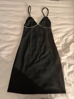 Svart satin klänning med strassdetalj - Helt ny Snygg svart klänning i satin med tunna axelband och strassdetalj längs bysten. Figurnära passform och kort modell som ger en elegant och trendig look. Perfekt för dig som gillar stilrena och glamorösa plagg.
