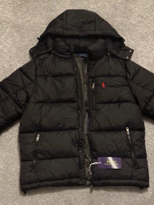 Svart pufferjacka från Polo Ralph Lauren - Snygg svart pufferjacka från Polo Ralph Lauren med huva och dragkedja framtill. Jackan har två sidofickor med dragkedja och en bröstficka. Ikoniska röda Polo-loggan broderad på bröstet. Perfekt för kalla vinterdagar.