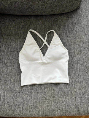 Vit sport-bh med korsade band - Säljer en vit sport-bh med smala axelband som korsas i ryggen. Bh:n har en djup v-ringning och är tillverkad i ett stretchigt, mjukt material som passar perfekt för träning eller yoga. Enkel och stilren design utan synligt märke.