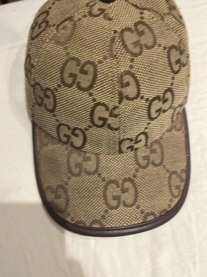 Gg keps - Säljer en beige keps från Gucci med det klassiska GG-mönstret i brunt över hela kepsen. Kepsen har böjd skärm med brun kant och är tillverkad i ett slitstarkt tygmaterial. Perfekt för dig som vill ha en stilren och lyxig accessoar.