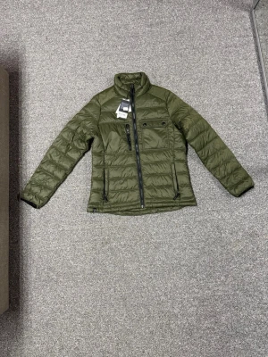 Olivgrön pufferjacka med dragkedja - Snygg olivgrön pufferjacka med hög krage och svart dragkedja framtill. Jackan har en bröstficka med knappar och två sidofickor med dragkedja. Tillverkad i glansigt, quiltat material som håller värmen under kalla dagar. Helt ny fick den i present och den passade inte med pris lapp. 