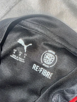 Svart och röd Puma sporttröja - Säljer en svart och röd sporttröja från Puma med MSC-logga på ärmen. Tillverkad i återvunnet material (RE:FIBRE) och har tryckta detaljer. Perfekt för träning eller match och har en modern, sportig look.