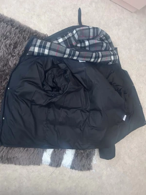 Svart pufferjacka från Burberry - Säljer en svart pufferjacka från Burberry med huva och dragkedja. Jackan har ett rutigt innerfoder i grått, vitt och rött samt en patch med texten 'BURBERRY LONDON ENGLAND' på ärmen. Perfekt för kalla vinterdagar och riktigt snygg streetstil. Jackan har ingen dragkedja men dragkedja medföljer storlek L och namnet som står burberry med vit text den är inte smutsig den är helt helt rent 