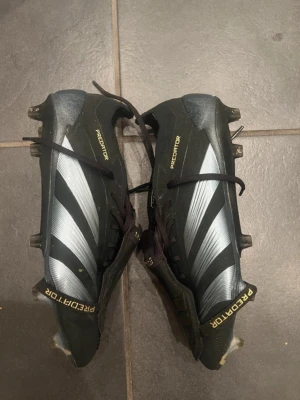 Adidas Predator svart/silver fotbollsskor - Snygga Adidas Predator fotbollsskor i svart med silvriga detaljer och klassiska tre ränder på sidan. Skorna har snörning, dobbar för bra grepp på planen och texten 'PREDATOR' i gult på häl och sula. Tillverkade i syntetmaterial för lätt känsla och bra bollkontroll.