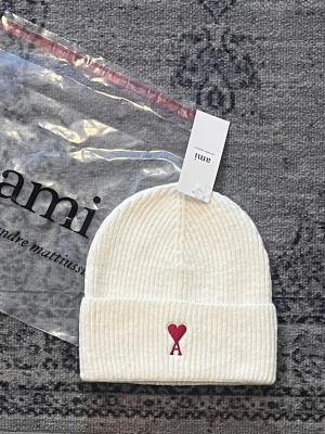 Vit ribbstickad mössa från Ami - Snygg vit ribbstickad mössa från Ami med uppvikt kant och röd broderad logga med hjärta och bokstaven A framtill. Klassisk passform och mjukt material som håller dig varm. Perfekt accessoar för kalla dagar.
