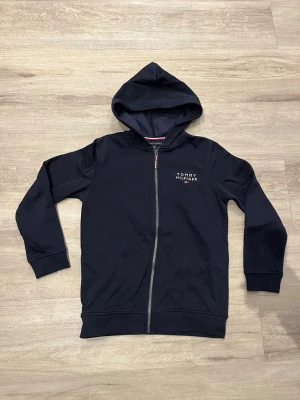 Mörkblått set från Tommy Hilfiger - Snyggt mörkblått set från Tommy Hilfiger med zip-hoodie och matchande byxor. Båda delarna har diskret logga på bröst och lår. Hoodien har dragkedja och fickor, byxorna har resår i midjan och muddar vid benslut. Perfekt för en avslappnad och sportig stil.
