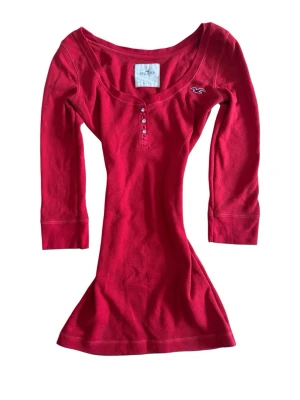 Vintage Röd ribbad Henley topp från Hollister - Röd ribbad topp från Hollister med trekvartsärm och knappar framtill. Toppen har en djupare rund halsringning och liten logga broderad på bröstet. Perfekt för en avslappnad och trendig look.