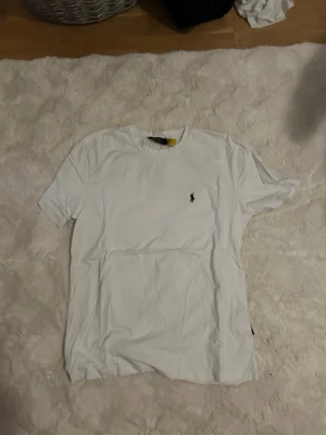 Vit t-shirt från Polo Ralph Lauren - Klassisk vit t-shirt från Polo Ralph Lauren med den ikoniska lilla broderade loggan på bröstet. T-shirten har rund hals och korta ärmar, perfekt för en clean och stilren look. Tillverkad i mjuk bomull för maximal komfort.