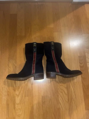 Tommy Hilfiger Whitney 2B boots storlek 39 – svart mocka - Säljer ett par snygga boots från Tommy Hilfiger, modell Whitney 2B. Storlek 39. Svarta i mocka med dekorativ röd detalj baktill och klassisk TH-logga. Dragkedja på insidan gör dem enkla att ta av och på. De är i bra skick. Skicka bara meddelande ifall ni undrar något mer.