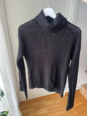 Svart stickad polotröja - Snygg svart stickad polotröja med grovstickat mönster och lång ärm. Tröjan har en hög krage och är perfekt för lager-på-lager-stil. Materialet känns mjukt och luftigt, vilket gör den både bekväm och trendig.