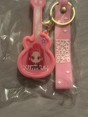 KPOP Demon Hunters rosa nyckelring - Säljer en söt nyckelring i form av en rosa gitarr med anime-inspirerad karaktär och texten 'KPOP DEMON HUNTERS' på ett matchande band. Nyckelringen har guldfärgad ring och karbinhake. Perfekt accessoar för K-pop fans! Som blinkar när man slår på den