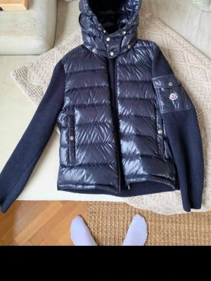 Moncler svart pufferjacka med stickade ärmar storlek m - Snygg svart pufferjacka från Moncler med glansig vadderad front och huva, samt stickade ärmar och rygg. Jackan har dragkedja, tryckknappar och en Moncler-logga på ärmen. Perfekt för kalla vinterdagar och riktigt trendig look. typ aldrig använd kemtvättades nyligen storlek M