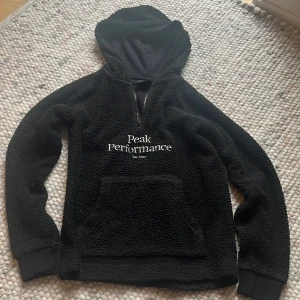 Svart fleecehoodie Peak Performance - Mysig svart hoodie från Peak Performance i fleece med broderad logga på bröstet. Tröjan har huva, halv dragkedja och stor känguruficka framtill. Perfekt för kyliga dagar och chill vibes.