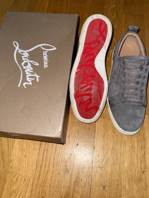 Gråa Christian Louboutin sneakers - Snygga gråa sneakers från Christian Louboutin i mocka med klassisk röd sula och vita kanter. Skorna har lågt skaft, matchande gråa skosnören och diskret branding på insidan. Perfekta för dig som vill ha något exklusivt och stilrent.