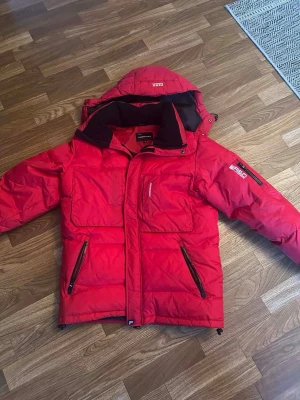 Röd dunjacka från Peak Performance - Snygg röd dunjacka från Peak Performance ( äldre model) väldigt bra skick då den aldrig ens är använd . Avtagbar huva och svarta detaljer. Flera stora fickor, broderad logga på ärmen och hög krage. Perfekt för kalla vinterdagar och riktigt varm.