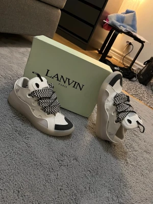 Lanvin vita sneakers med mönstrade snören - Lanvin sneakers i vitt läder med svarta detaljer och breda, svartvita mönstrade snören. Snygg chunky sula och perforerade partier på tån. Klassisk low-top modell med unik design och logga på sidan. Perfekta för dig som vill sticka ut med dina skor. Förmodligen inte äkta.