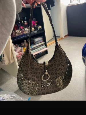 Brun axelremsväska - Snygg brun handväska med klassiskt G-mönster och detaljer i krokopräglat konstläder. Väskan har en rundad form, dekorativ metallring framtill och dragkedja på baksidan. Perfekt storlek för det viktigaste och trendig axelrem. Köptes utomlands.