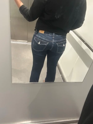 Mörkblå bootcut jeans med kontrastsömmar - Säljer ett par mörkblå bootcut jeans med vita kontrastsömmar och snygga fickdetaljer bak. Jeansen har normal midja och fickor med lock och knapp bak. Perfekta för dig som gillar klassisk men ändå trendig stil.