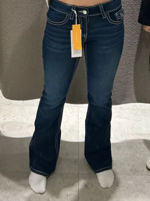 Mörkblå bootcut jeans low waist - Säljer ett par mörkblå bootcut jeans från Gina med vita kontrastsömmar och låg midja. Jeansen har klassiska fem fickor, knappgylf och dekorativ knapp på bakfickan. Perfekta för en trendig och avslappnad look. Skriv för fler bilder eller frågor💞