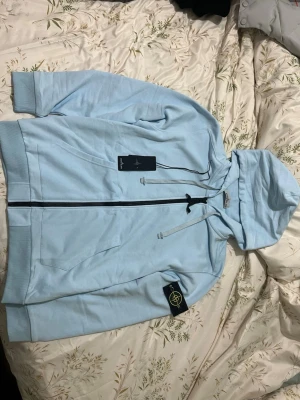 Ljusblå hoodie från Stone Island - Snygg ljusblå hoodie från Stone Island med dragkedja framtill och klassisk logopatch på vänster ärm. Hoodien har justerbar huva med snören och två fickor framtill. Tillverkad i mjuk bomull för skön känsla och stilren look.