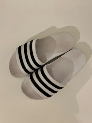 Vita Adidas tofflor - Säljer ett par vita adidas tofflor i strl 38. De är använda men ändå i bra skick. Skriv gärna vid frågor!🩷