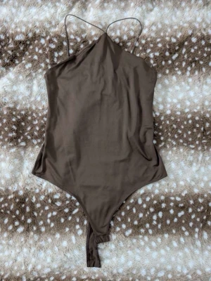 Brun body med tunna axelband - Säljer en stilren brun body med tunna axelband och tryckknappar i grenen. Bodyn har en enkel, slät design och är tillverkad i ett mjukt stretchigt material som sitter snyggt på kroppen. Perfekt att matcha med jeans eller kjol för en trendig look.