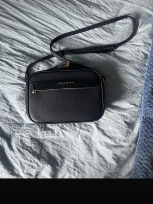 Mörkblå axelväska från Laura Ashley - Snygg mörkblå axelväska från Laura Ashley med guldfärgade detaljer och justerbar axelrem. Väskan har en stilren, rektangulär form och är tillverkad i ett tåligt konstläder. Framtill finns en ficka med dragkedja och diskret logga i guld. Perfekt för att bära det viktigaste.