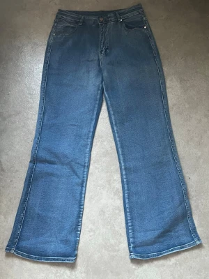 Blåa bootcut jeans med hög midja - Ett par klassiska blåa bootcut jeans med hög midja och fem fickor. Jeansen har en lätt utsvängd passform nedtill och är sydda i ett mjukt denimtyg. Perfekta för en avslappnad och trendig look.