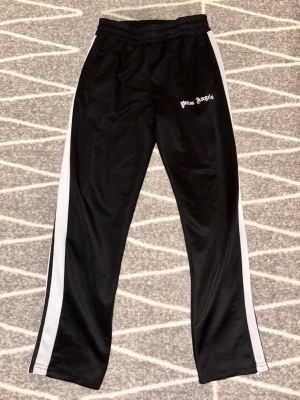 Palm Angels svarta mjukisbyxor - Palm Angels mjukisbyxor i svart med vita sidoränder och logga tryckt på vänster sida. Byxorna har resår i midjan och raka ben. Tillverkade i ett glansigt, sportigt material som känns riktigt skönt och ger en clean streetwear-look.