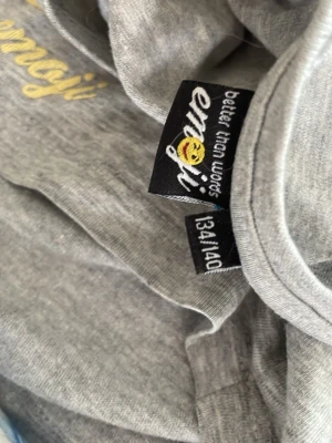 Grå t-shirt från Emoji - Säljer en enkel och stilren grå t-shirt från Emoji med gul textdetalj på bröstet. T-shirten har korta ärmar och är gjord i mjuk bomull, perfekt för en avslappnad look.