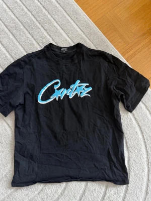 Svart Corteiz t-shirt med blått tryck - Säljer en svart t-shirt från Corteiz med ett stort blått och vitt grafiskt tryck på bröstet. Klassisk rund hals och korta ärmar. Perfekt för dig som gillar streetwear och vill sticka ut med en snygg logga framtill.