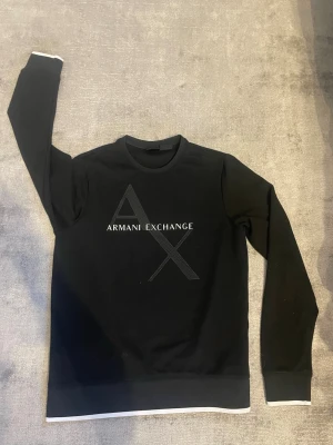 Svart sweatshirt från Armani Exchange - Svart sweatshirt med stor AX-logga och texten 'Armani Exchange' på bröstet. Tröjan har rund hals, långa ärmar och vita detaljer vid ärmslut och nederkant. Perfekt för en clean och stilren look. Nypris: ca 900kr. Mitt pris: 350kr. Storlek M