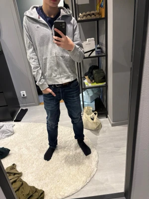 Grå zip hoodie från Tommy Hilfiger - Säljer en stilren grå hoodie från Tommy Hilfiger med dragkedja framtill och klassisk huva. Hoodien har två fickor fram och diskret logga på bröstet. Perfekt till jeans eller joggers för en chill look.