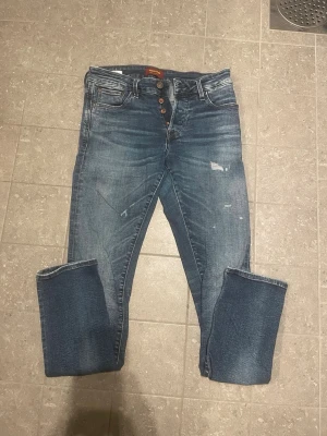 Blå jeans med knappar från Jack & Jones - Säljer ett par blå jeans från Jack and jones med knäppning framtill och slitna detaljer. Jeansen har fem fickor, raka ben och uppvikta benslut. Klassisk tvättad denim med coola slitningar och Nudie-lapp bak i midjan.