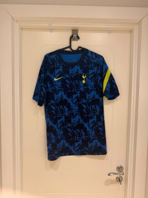 Tottenham Hotspur blå tränings t-shirt - Snygg blå tränings t-shirt från Nike med Tottenham Hotspur-logga på bröstet och gul Nike swoosh. Tröjan har ett unikt blått mönster och en gul detalj på ärmen. Perfekt för fotbollsträning eller gymmet. Tillverkad i lätt och ventilerande material.                           Modellen är 185cm lång och väger 82kg.