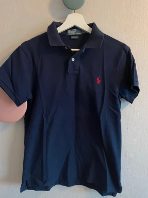 Marinblå piké från Ralph Lauren - Klassisk marinblå pikétröja från Ralph Lauren med röd broderad logga på bröstet. Slim fit-modell med korta ärmar, krage och knappar framtill. Tillverkad i mjuk bomull som känns skön mot huden. Storleken är m men den sitter som s-m