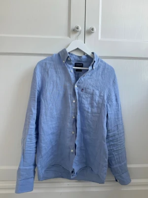 Ljusblå linneskjorta från Lexington - Säljer en stilren ljusblå skjorta från Lexington i svalt linne. Skjortan har klassisk krage, bröstficka och vita knappar framtill. Perfekt till sommaren och enkel att matcha med jeans eller shorts.
