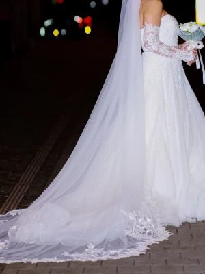 Ivory brudklänning med spets och släp från Lilly - En vacker brudklänning i stl 36/38 med intrikata spetsdetaljer och pärlbroderier över hela klänningen. Klänningen har en off-shoulder design med långa ärmar i spets och en elegant släp. Ett glittrigt bälte markerar midjan och ger en fin silhuett. Klänningen är i en klassisk A-linjeform som faller vackert. Obs! Kemtvättad Klänningen är köpt hos Joliebridal i Malmö för 19,999kr Special designade armar 1500kr Brudslöja 2000kr Bälte 500kr Totalt 23,999kr och vill ge den till en annan lycklig kvinna 