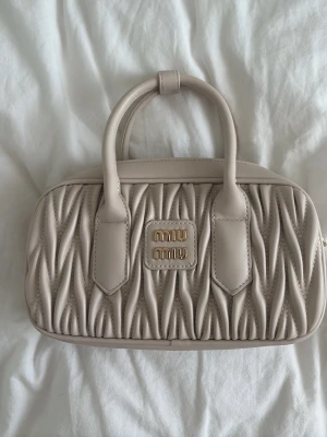 Beige handväska  - Säljer en beige handväska i mjukt quiltat läder med två handtag och guldfärgad logga framtill. Väskan har en unik vågig struktur och klassisk rektangulär form. Perfekt för dig som gillar stilrena och exklusiva accessoarer.