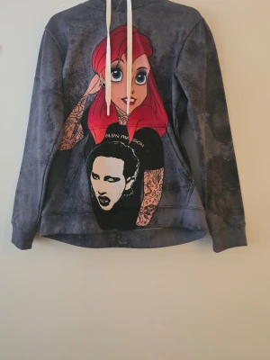 Grå hoodie med tryck Ariel Manson, XS - Unik grå hoodie med coolt tryck på ryggen av en tatuerad person med rött hår och ett ansikte på tröjan. Stora vita dragsnören i huvan och texten 'ARIEL MANSON' på ryggen. Mjuk och bekväm, perfekt för dig som gillar streetwear och statement-plagg.