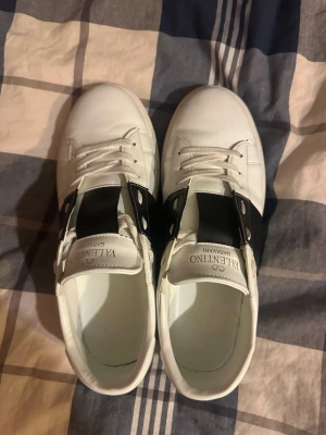 Valentino vita sneakers med svart detalj - Snygga vita sneakers från Valentino Garavani med bred svart läderdetalj över snörningen och nitar längs hälen. Klassisk låg modell med vita skosnören och diskret logga på plösen. Tillverkade i slätt läder för en clean och modern look.