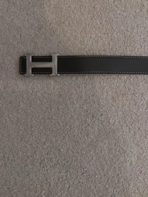 Svart skärp med H-spänne - Snyggt svart bälte med ett stort H-format metallspänne och vita kontrastsömmar. Perfekt accessoar för att lyfta din outfit med en stilren och klassisk look. Bältet är gjort i skinn och har en exklusiv känsla.