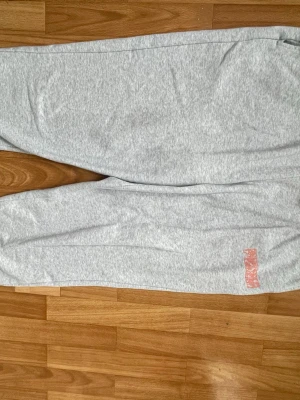 Gråa mjukisbyxor från Puma - Snygga ljusgrå mjukisbyxor från Puma med resår i midjan och dragsko för skön passform. Byxorna har ett diskret Puma-tryck i rött på benet och är tillverkade i mjukt bomullsmaterial. Perfekta för chill dagar eller träning. Tveka inte på att höra av dig om du har några frågor!