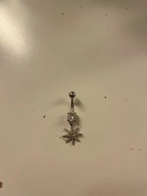 Navelpiercing med stjärnhänge - Snygg navelpiercing i silverfärgad metall med en gnistrande klar sten och ett hängande stjärnformat smycke dekorerat med små stenar. Perfekt accessoar för att ge din look lite extra bling. Använd 1 gång men kokad och rengjord med handsprit. TROR DE ÄR ÄKTA SILVER