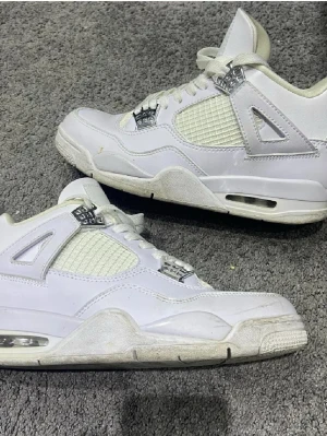 Vita Air Jordan 4 Retro sneakers - Säljer ett par klassiska vita Air Jordan 4 Retro sneakers med meshpaneler och detaljer i silver. Skorna har synlig Air-sula, ikonisk Jumpman-logga på hälen och coola plastdetaljer vid snörningen. Perfekta för dig som gillar streetwear och vill sticka ut.